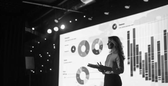 Woman giving a presentation with a large screen of statistics projected behind her.